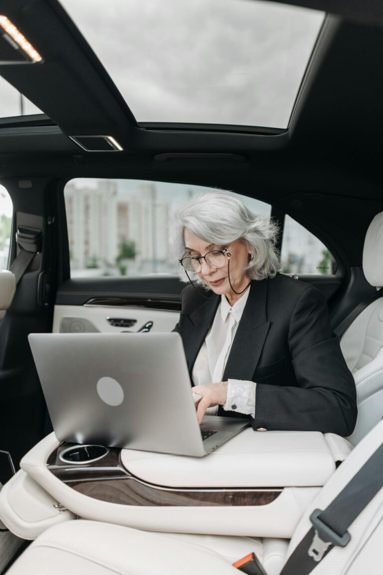 A senior businesswoman with gray hair using a laptop in a luxury car interior.